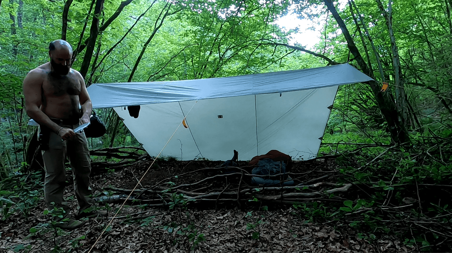 Man with knife in front of tarp set up in the forest